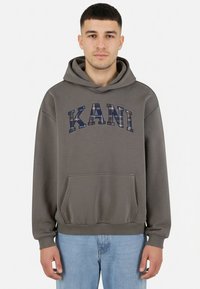 Noor mees, kes kannab halli kapuutsiga dressipluusi, millel on heledast ruudulisest mustrist "KANI" tekst, helerohelised teksad, seisab valge taustaga ees.