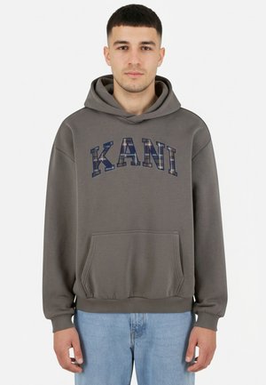 Jonge man die een grijze hoodie draagt met de tekst "KANI" in ruitpatroon, lichtblauwe spijkerbroek, staand tegen een effen witte achtergrond.