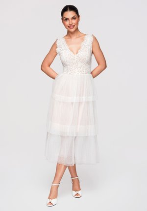 Femme portant une robe en dentelle blanche sans manches avec une jupe en tulle et des talons blancs ouverts à la cheville avec bride, debout les mains dans les poches, souriante.