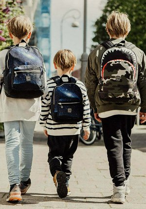Drie kinderen lopen weg, elk met een unieke rugzak: een met een sterrenstelselprint, een zwart-wit gestreept en een camouflageontwerp met rode accenten.