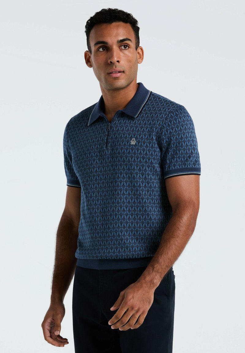La chemise polo à motifs bleus présente un col zippé, des manches courtes et des finitions contrastées en marine. Le tissu a une apparence texturée et tissée.
