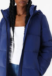 Jachetă puffer albastră cu glugă, realizată dintr-un material neted și lucios. Dispune de un fermoar frontal, accente elastice și secțiuni quilted orizontale.