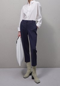 Camisa blanca abotonada, pantalones sastre azul marino, calcetines verdes con zapatos de tacón bloque, sosteniendo un bolso de mano blanco frente a un fondo texturizado.
