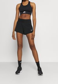 Sostén deportivo negro con el logo de Nike Air, combinado con pantalones cortos deportivos negros con una cinturilla oculta. Zapatillas negras y calcetines completan el conjunto.