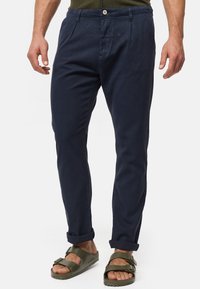 Navy chinos made of cotton with a smooth texture, featuring front pleats, front and back pockets, and rolled cuffs, paired with green sandals.