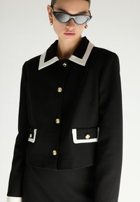 Schwarze kurze Jacke mit weißem Kragen; ausgestattet mit goldenen Knöpfen und Taschen mit weißem Rand. Glattes Material mit maßgeschneiderten Designdetails.