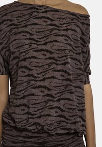 Μωβ τοπ με εκτεθειμένους ώμους και μαύρα αφηρημένα κυματιστά σχέδια, φορεμένο στο πάνω μέρος του σώματος, που δείχνει έναν γυμνό ώμο και κοντά μανίκια.