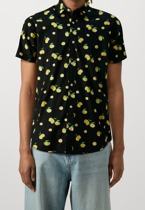 Chemise noire à manches courtes avec boutons, ornée d'un motif de citrons jaunes et de points, portée avec un jean bleu clair par une personne debout.