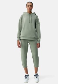 Felpa verde chiaro e pantaloni sportivi cropped in morbido materiale, dotati di cappuccio, tasca a marsupio e orlo a coste, abbinati a sneakers bianche.
