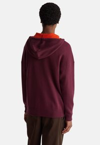 Sudadera con capucha color burdeos de corte relajado, con un cuello naranja en contraste y puños acanalados. Textura suave y diseño casual.