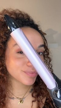 Roze krultang met glanzende zwarte accenten, voorzien van een cilindervormige barrel en een textuurrijke handgreep. Close-up weergave naast het gezicht van een persoon.