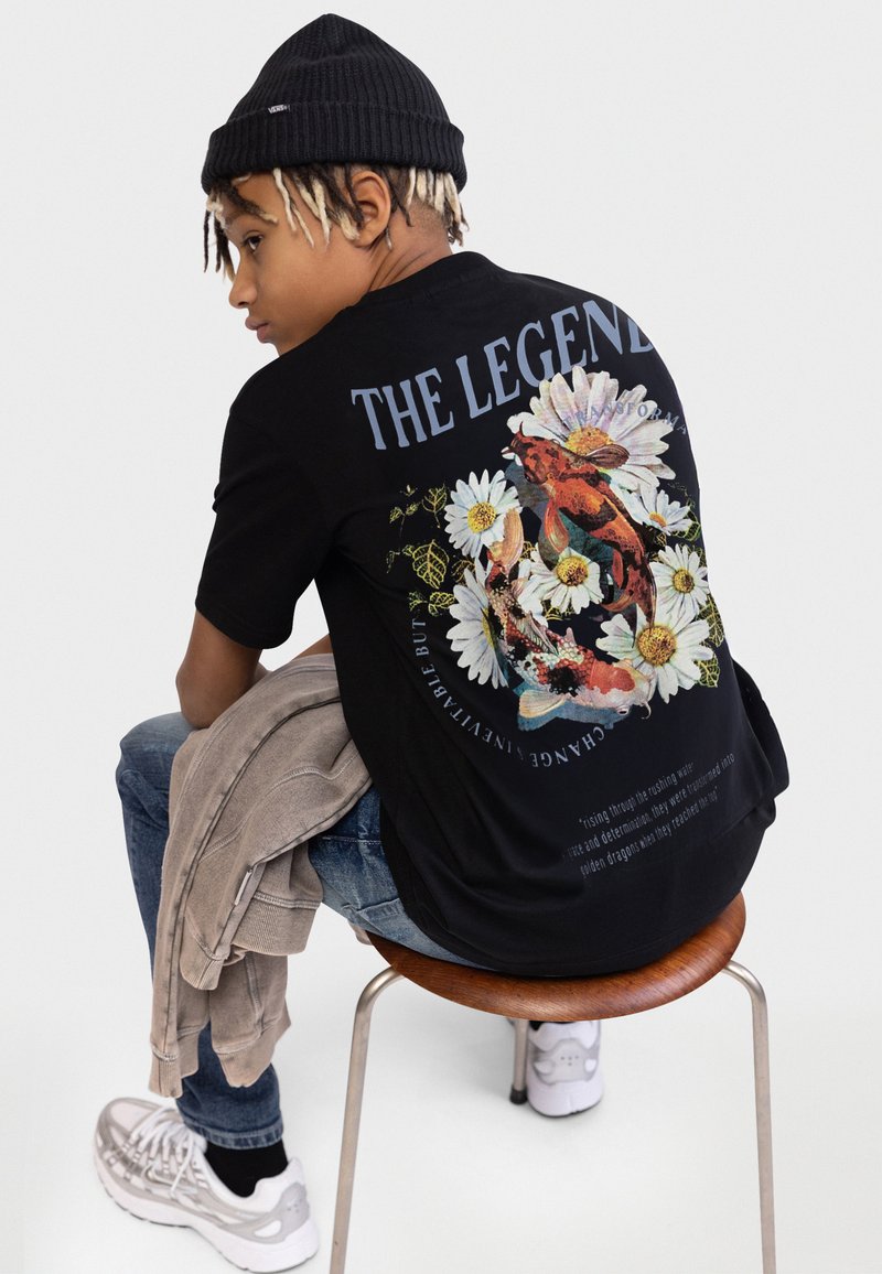 Jongere met een zwarte muts en een zwart T-shirt met een bloem- en koi-visontwerp, zittend op een houten kruk en een beige jas vasthoudend.