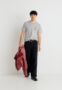 Νεαρός άντρας περπατάει, φορώντας γκρι μπλουζάκι, μαύρο παντελόνι, μαύρα παπούτσια, κρατώντας ένα διπλωμένο κόκκινο μπουφάν στο δεξί του χέρι πάνω σε απλό λευκό φόντο.