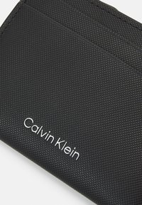 Portafoglio nero con texture, dotato di scomparti per carte e un logo "Calvin Klein" in argento impressionato sulla superficie. Numerosi vani per una migliore organizzazione.
