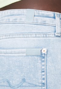 Un jean en denim bleu clair présente une surface texturée, une poche arrière avec une étiquette en cuir bleu clair embossée du texte « for all mankind », et des accessoires en argent.