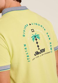 Polo jaune avec un col contrasté et des graphiques brodés incluant des palmiers et des éléments de texte au dos.