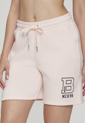 Light pink drawstring shorts with pockets worn by a person, featuring black "B Bench MCR'89" text on the lower left leg.