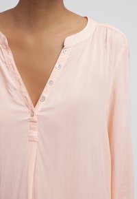 Blouse de couleur pêche avec un col en V et des manches longues, présentant des accents de boutons le long de la patte de boutonnage et une texture de tissu lisse et léger.