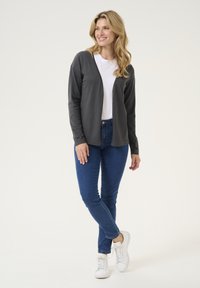 Šedý cardigan přes bílou košili, modré džíny a bílé tenisky. Má dlouhé rukávy a otevřený design. Hladká textura látky.