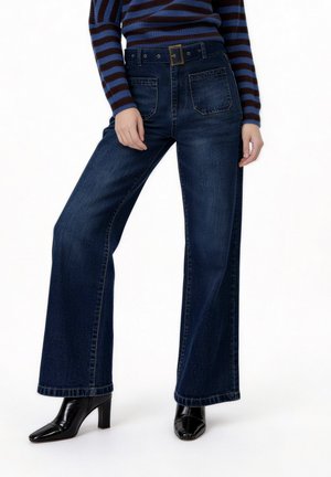 Άτομο που φοράει σκούρο μπλε τζιν με φαρδιά παντελόνια, εμπρός με τσέπες κεντήματος, και μαύρα μποτάκια με τακούνι, σε συνδυασμό με ένα μπλε και καφέ ριγέ πουλόβερ.