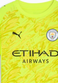 Maglia gialla brillante con motivi astratti, logo Puma in nero, stemma del Manchester City e "ETIHAD AIRWAYS" stampato in lettere nere in grassetto.