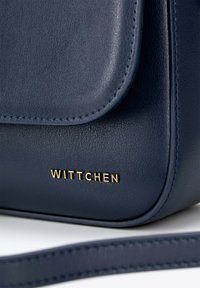 Marineblaue Lederhandtasche mit glatter Textur, gekennzeichnet durch einen geschwungenen Deckel, das goldene "WITTCHEN"-Markenlogo und einen abnehmbaren Gurt.