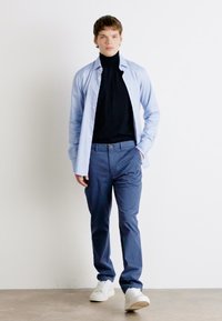Lys blå skjorte med knapper lagd over en svart turtleneck, kombinert med marineblå bukser og hvite joggesko. Enkelt, moderne design og farger.