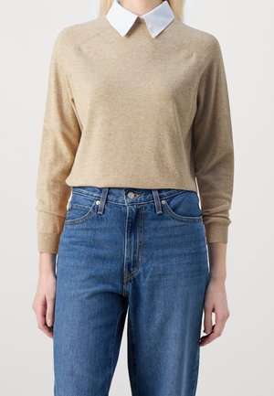 Personne portant un pull beige à manches longues sur une chemise blanche à col, associée à un jean en denim bleu taille haute, sur un fond uni.