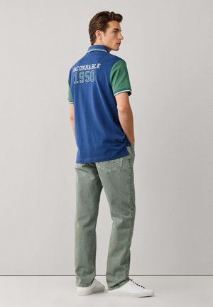 Hombre de espaldas vestido con una camisa polo azul y verde con "FACONNABLE 1950" en la parte trasera, pantalones verdes y zapatillas blancas.