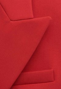 Gros plan sur le revers et la poche d'un blazer rouge, mettant en valeur le tissu texturé et les détails de couture ajustée.