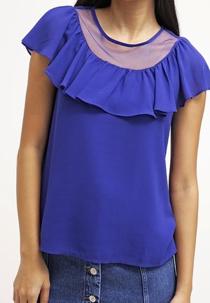 Koningblauwe blouse met korte ruches aan de mouwen, een transparant paneel bij de halslijn en een gladde textuur. Draagt bij een denim rok.