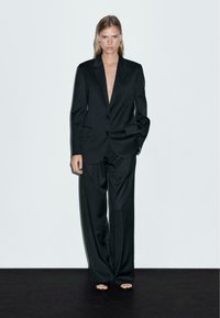 Svart skräddarsydd kostym med en djup V-ringad blazer, vida byxor och minimalistiska svarta sandaler. Släthet tyg med en elegant finish.