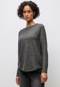 Kvinde med kort krøllet hår iført en mørkegrå langærmet sweater og sorte jeans, stående foran en ensartet lys baggrund.