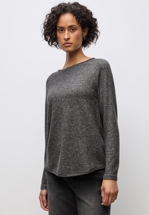 Kvinde med kort krøllet hår iført en mørkegrå langærmet sweater og sorte jeans, stående foran en ensartet lys baggrund.