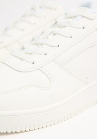 Zapatillas de cuero blancas con un acabado liso, que presentan cordones blancos planos, detalles de costura sutiles y una suela de goma texturizada.