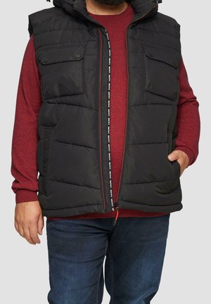 Homme portant une veste matelassée noire sur un pull rouge et un jean bleu, une main dans la poche de la veste et une barbe visible.