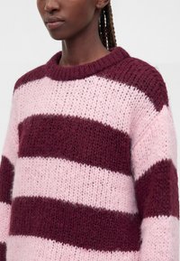 Femme portant un pull en maille duveteuse avec de larges rayures horizontales bordeaux et rose clair, les cheveux tressés visibles à l'arrière.