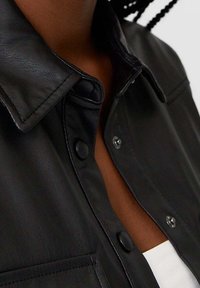 Chemise en cuir noir avec un col, dotée de boutons-pression et d'une poche poitrine. Texture lisse et finition mate. Gros plan sur les coutures détaillées.
