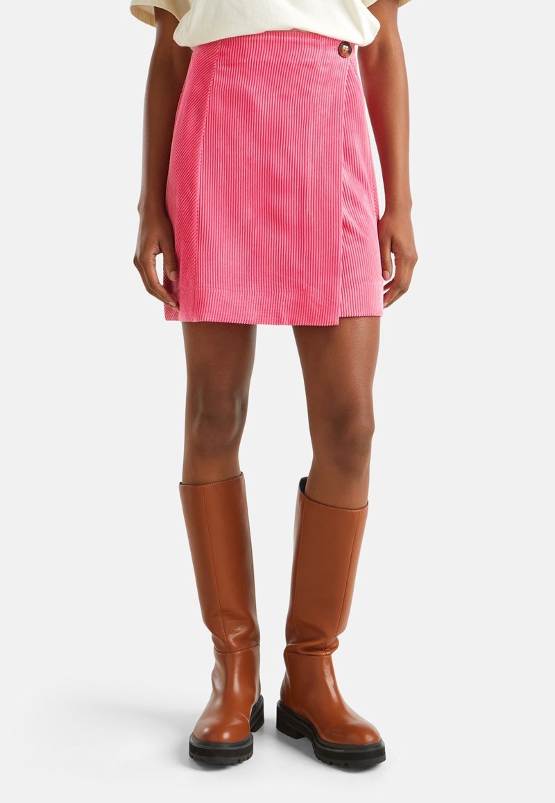 Woman wearing a bright pink corduroy mini skirt and knee-high brown leather boots, standing against a white background.