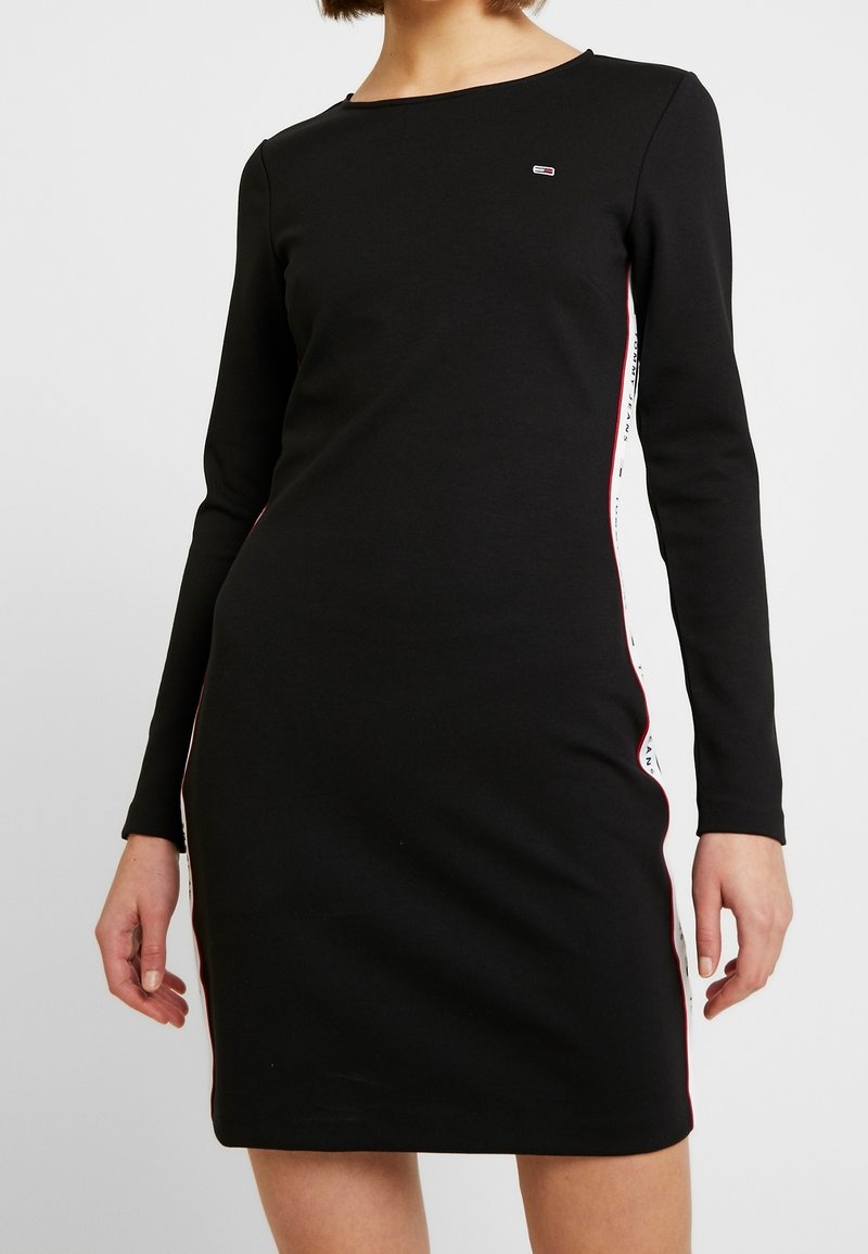 Robe noire ajustée à manches longues avec des rayures latérales blanches et rouges et un petit logo rectangulaire sur le haut de la poitrine.