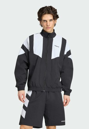 Junger Mann trägt eine schwarz-weiße Adidas-Trainingsjacke und passende Shorts, steht vor einem schlichten hellen Hintergrund.