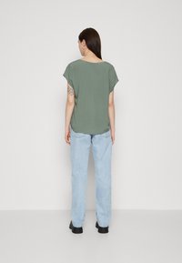Blusa corta verde oliva con maniche a cappuccio, abbinata a jeans straight-leg blu chiaro. Stivali neri e tatuaggio di ragnatela sul braccio visibili.