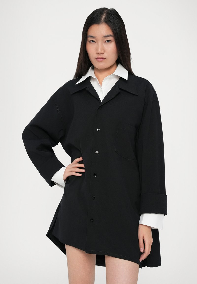 Ung kvinde med langt mørkt hår iført en oversized sort skjorte med knapper over en hvid kravebluse, poserer med den ene hånd på hoften.