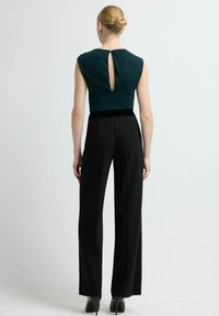 Donkergroene mouwloze top met een sleutelgatdetail aan de achterkant, gecombineerd met zwarte wijde broek met hoge taille en zwarte hoge hakken.