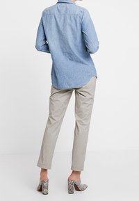 Personne portant une chemise en denim clair à manches longues, un pantalon beige et des chaussures à talons en motif serpent, se tenant devant un fond blanc.