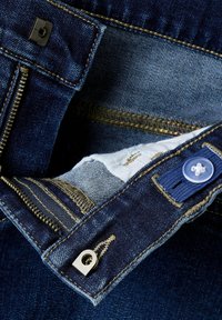 Interior de unos pantalones vaqueros de mezclilla oscuros mostrando un zipper, cierre de presión de metal y botón azul. Presenta costuras doradas y un forro de mezclilla clara.