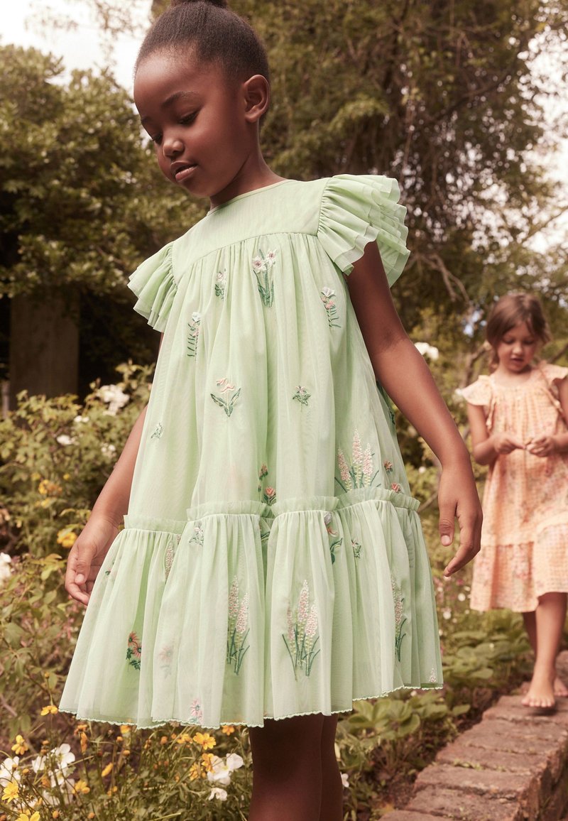 Vaaleanvihreä mekko, jossa on puffihihat, kukkakirjontaa, röyhelökerroksia ja löysä istuvuus. Kaksi lasta puutarha-asetelmassa.