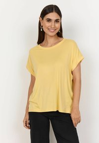 Gul ribbet T-shirt med korte ærmer og en løs pasform. Kombineret med sorte bukser og båret af en person med glat hår og øreringe.