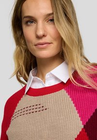 Strickpullover mit roten, rosa und beigen geometrischen Mustern, mit geripptem Ausschnitt und Schulterakzenten. Glatte Textur mit subtilen Details.