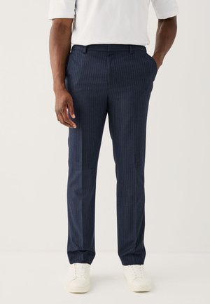 Muž nosiace námornícky modré nohavice s tenkými pruhmi, ruky v vreckách a biele tenisky, stojaci pred jednoliatym svetlým pozadím.